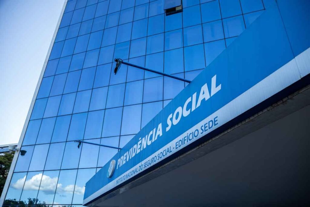 Fachada de edifício moderno com revestimento de vidro espelhado azul. Em primeiro plano, uma grande placa azul exibe os dizeres “PREVIDÊNCIA SOCIAL” e, abaixo, “INSTITUTO NACIONAL DO SEGURO SOCIAL - EDIFÍCIO SEDE”. O prédio reflete o céu e nuvens nas janelas, e a foto foi capturada em ângulo inclinado, destacando a altura da construção.