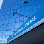 Fachada de edifício moderno com revestimento de vidro espelhado azul. Em primeiro plano, uma grande placa azul exibe os dizeres “PREVIDÊNCIA SOCIAL” e, abaixo, “INSTITUTO NACIONAL DO SEGURO SOCIAL - EDIFÍCIO SEDE”. O prédio reflete o céu e nuvens nas janelas, e a foto foi capturada em ângulo inclinado, destacando a altura da construção.