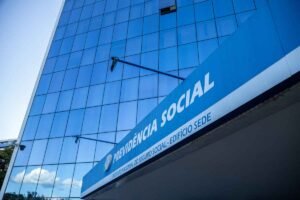 Fachada de edifício moderno com revestimento de vidro espelhado azul. Em primeiro plano, uma grande placa azul exibe os dizeres “PREVIDÊNCIA SOCIAL” e, abaixo, “INSTITUTO NACIONAL DO SEGURO SOCIAL - EDIFÍCIO SEDE”. O prédio reflete o céu e nuvens nas janelas, e a foto foi capturada em ângulo inclinado, destacando a altura da construção.